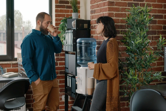 BulkBar - Fontaines à eau - 2 fontaines a eau au bureau - Deux collègues en entreprise se tiennent devant une fontaine à eau à bonbonne, l’un buvant dans un verre, l’autre discutant
