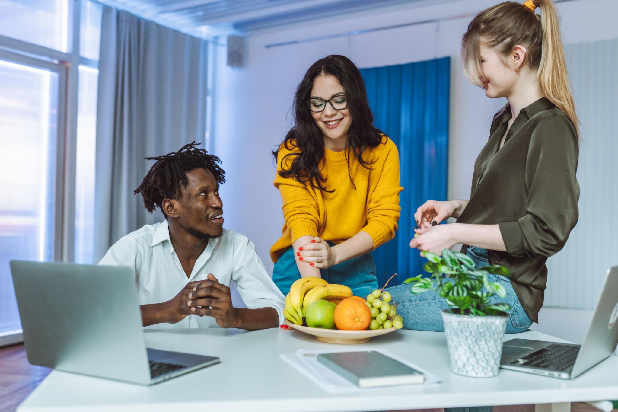 free food op kantoor - Drie collega's praten vrolijk met elkaar in een modern kantoor rond een schaal met fruit